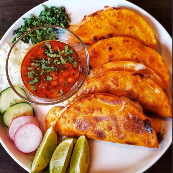 A plate of golden-brown birria tacos with a side of consommé, limes, radishes, cucumbers, and cilantro.