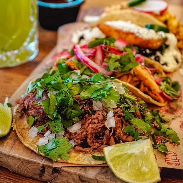 Close-up of three Mexican tacos with various fillings, cilantro, and lime wedges on a wooden board.