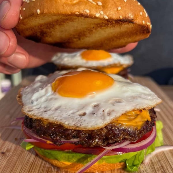 A hand places a sesame seed bun on a burger with a fried egg, cheese, and vegetables.