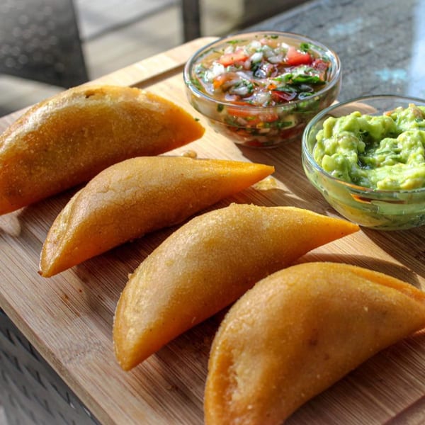 Four golden-brown empanadas on a wooden board, with bowls of salsa and guacamole.