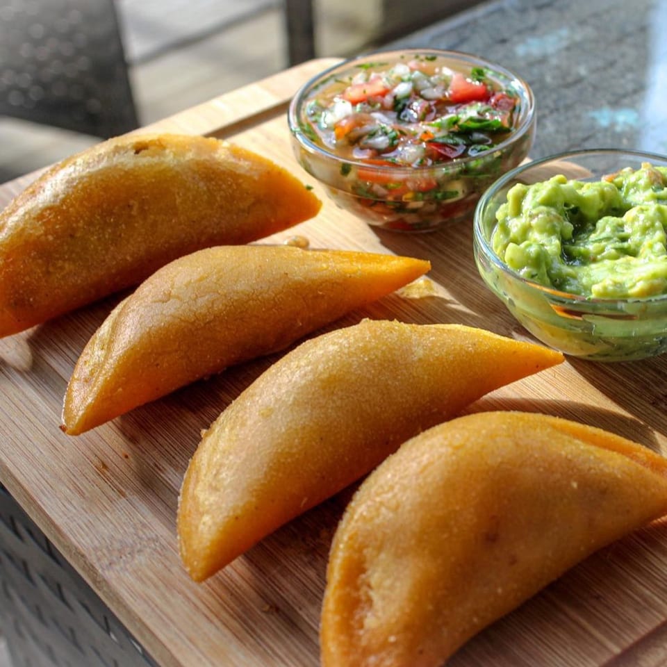 Four golden-brown empanadas on a wooden board, with bowls of salsa and guacamole.