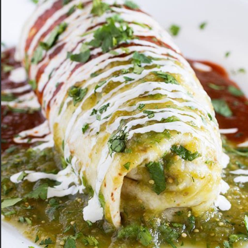 A large Mexican burrito covered in green and red sauces, drizzled with sour cream and cilantro.