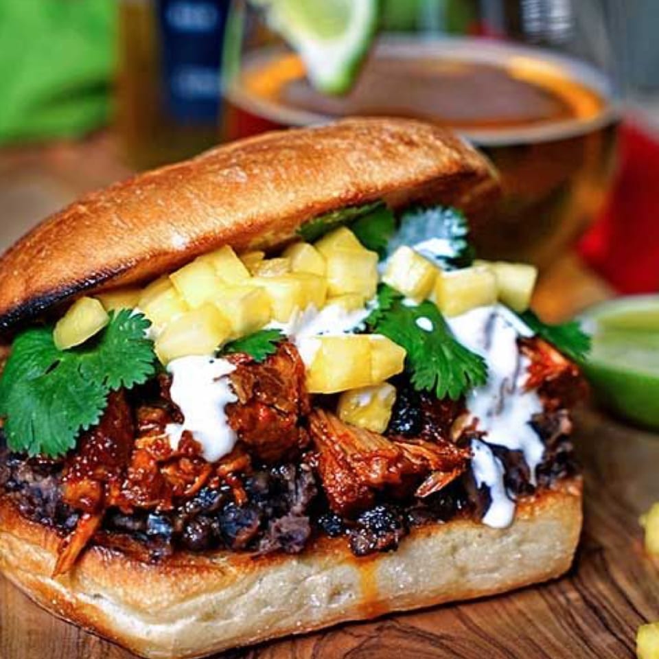 Mexican torta with pulled pork, black beans, pineapple, cilantro, and crema, with a drink in the background.