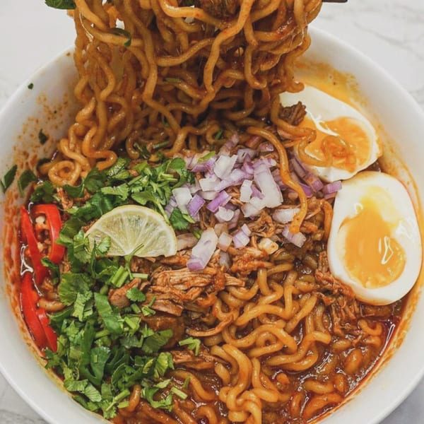 Spicy ramen with pulled pork, soft-boiled eggs, cilantro, red onion, and lime in a white bowl.
